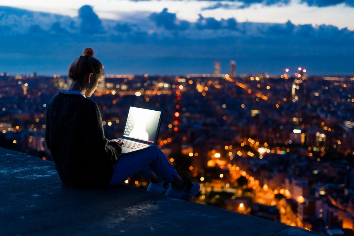 Chica joven viajera en lo alto de un mirador usando un ordenador portátil con ciudad iluminada de fondo en el atardecer   Young